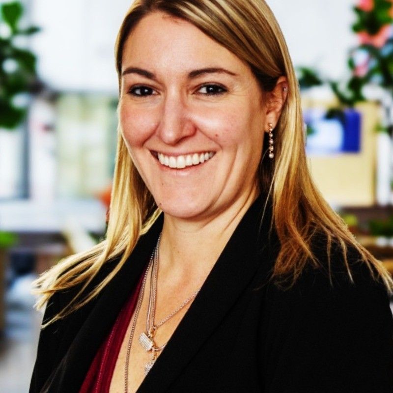 Sarah Malchow. Blonde woman smiles, wearing a black blazer, burgundy top, and silver necklaces.