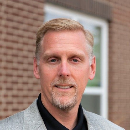 Todd Rodden. Man with fair hair and goatee in a gray blazer smiles in front of a brick wall.