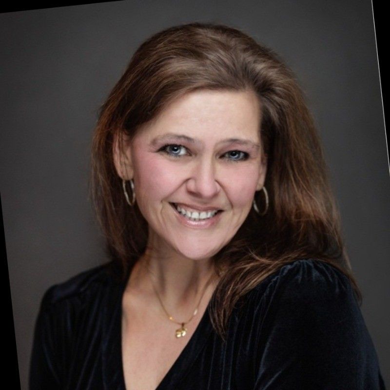Jennifer M Wheeler. Woman with long brown hair smiling, wearing a black top and gold jewelry against a gray background.