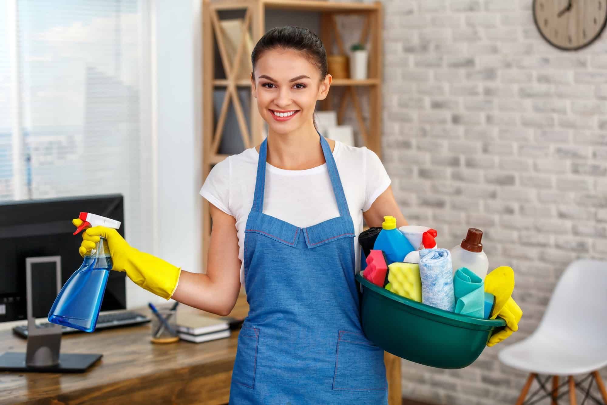 A woman is holding a bucket of cleaning supplies and a spray bottle.
