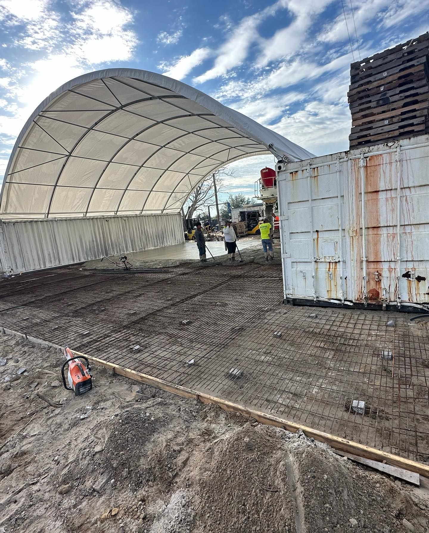 Construction site with arched white tent, concrete, and shipping container. Workers visible.