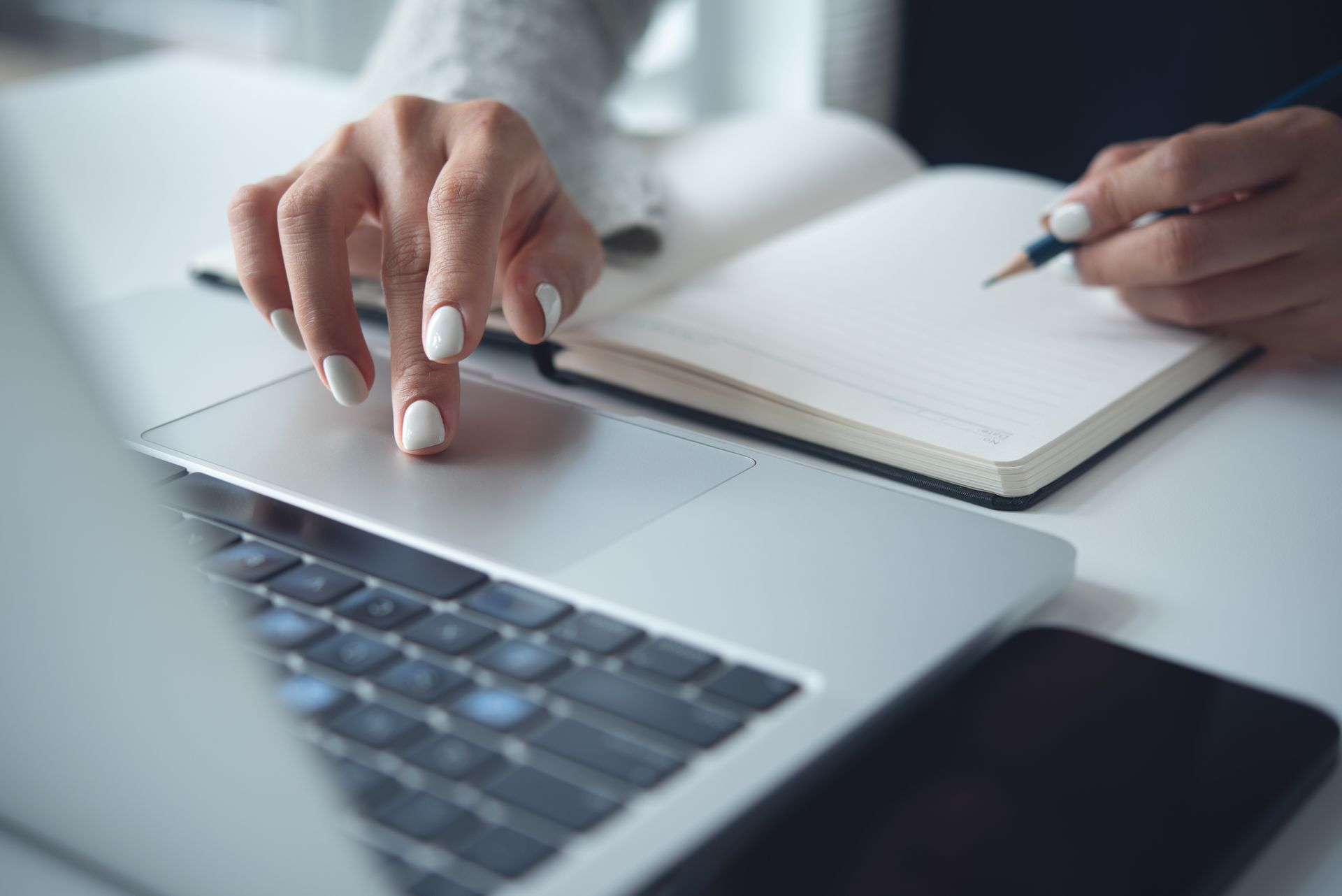 Person's hands using a laptop touchpad, with a notebook and pen open beside it.