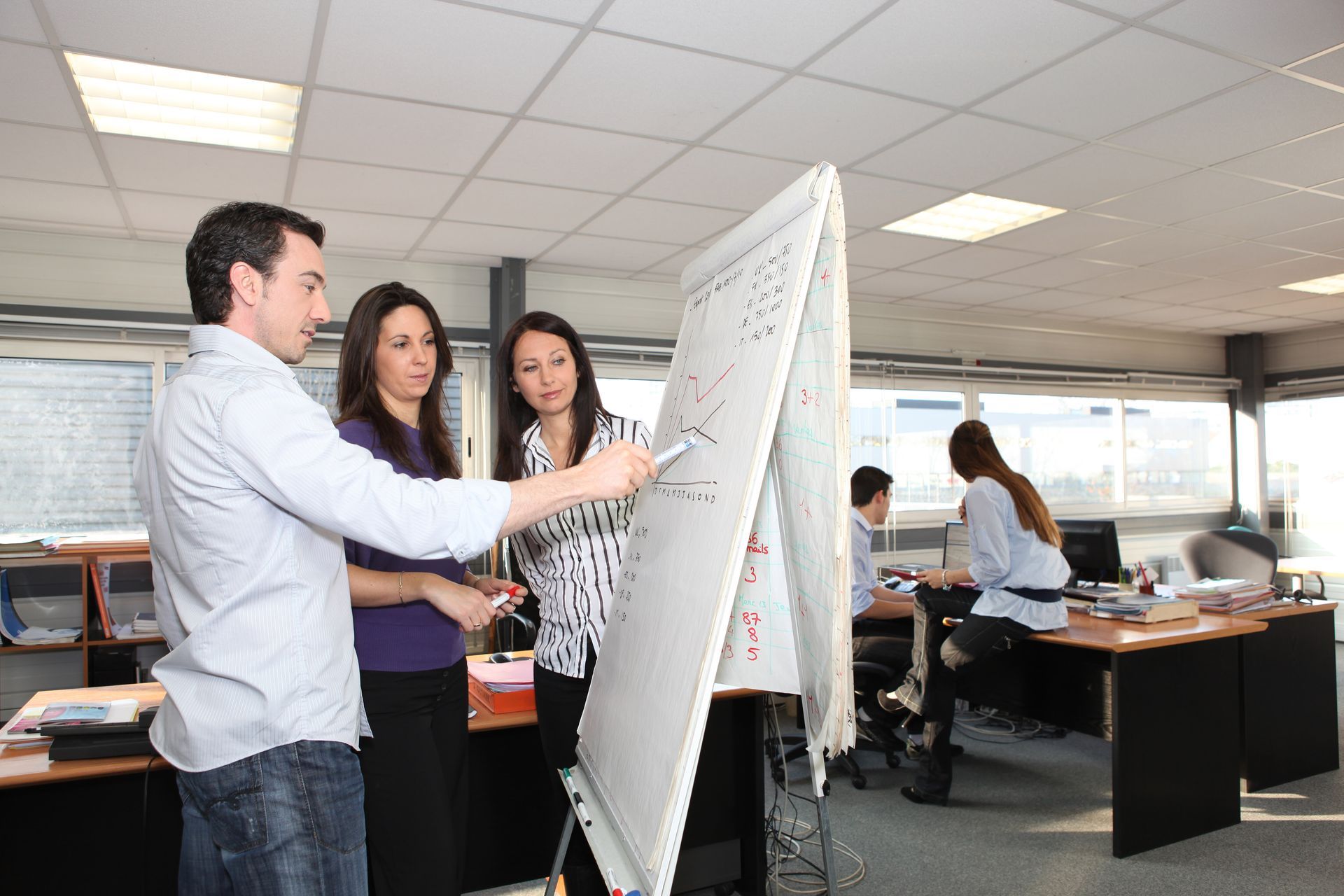 Three people in an office, one pointing at a whiteboard as two others look on, another working at a desk.