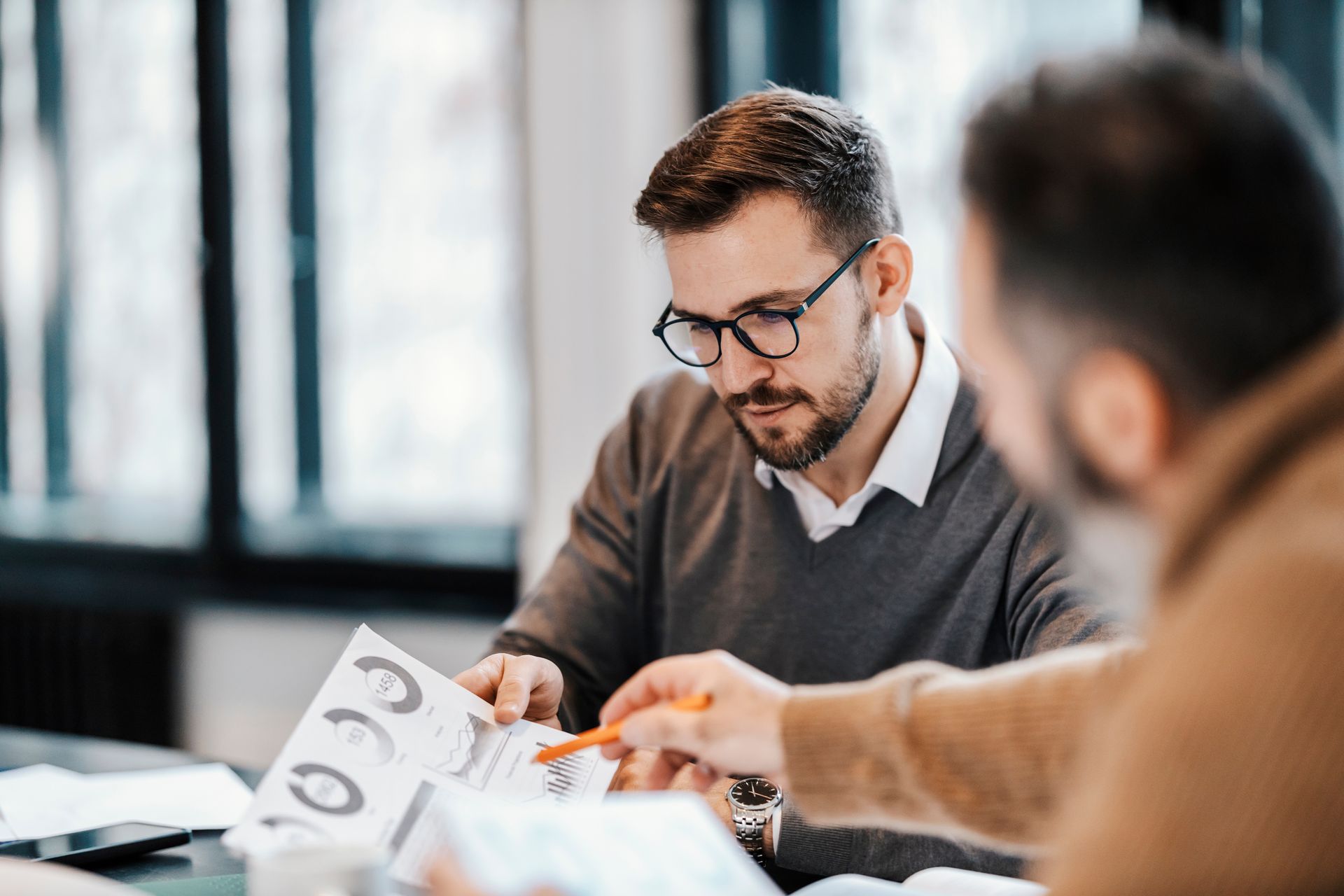 Man in glasses reviews a document with another man at a table. Office setting.