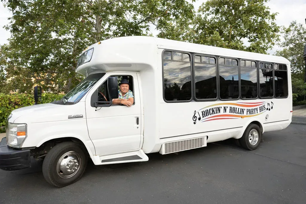 White passenger van parked on a road, with a driver visible and trees in the background