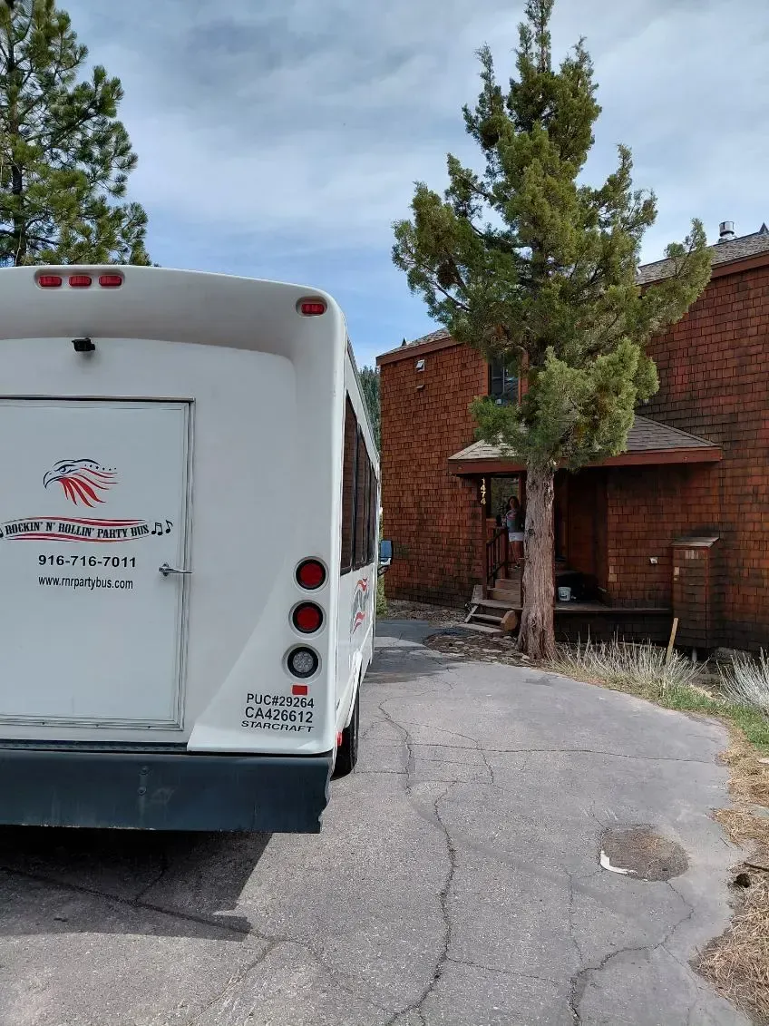 White bus parked on a cracked driveway beside red-brick buildings and pine trees under a cloudy sky