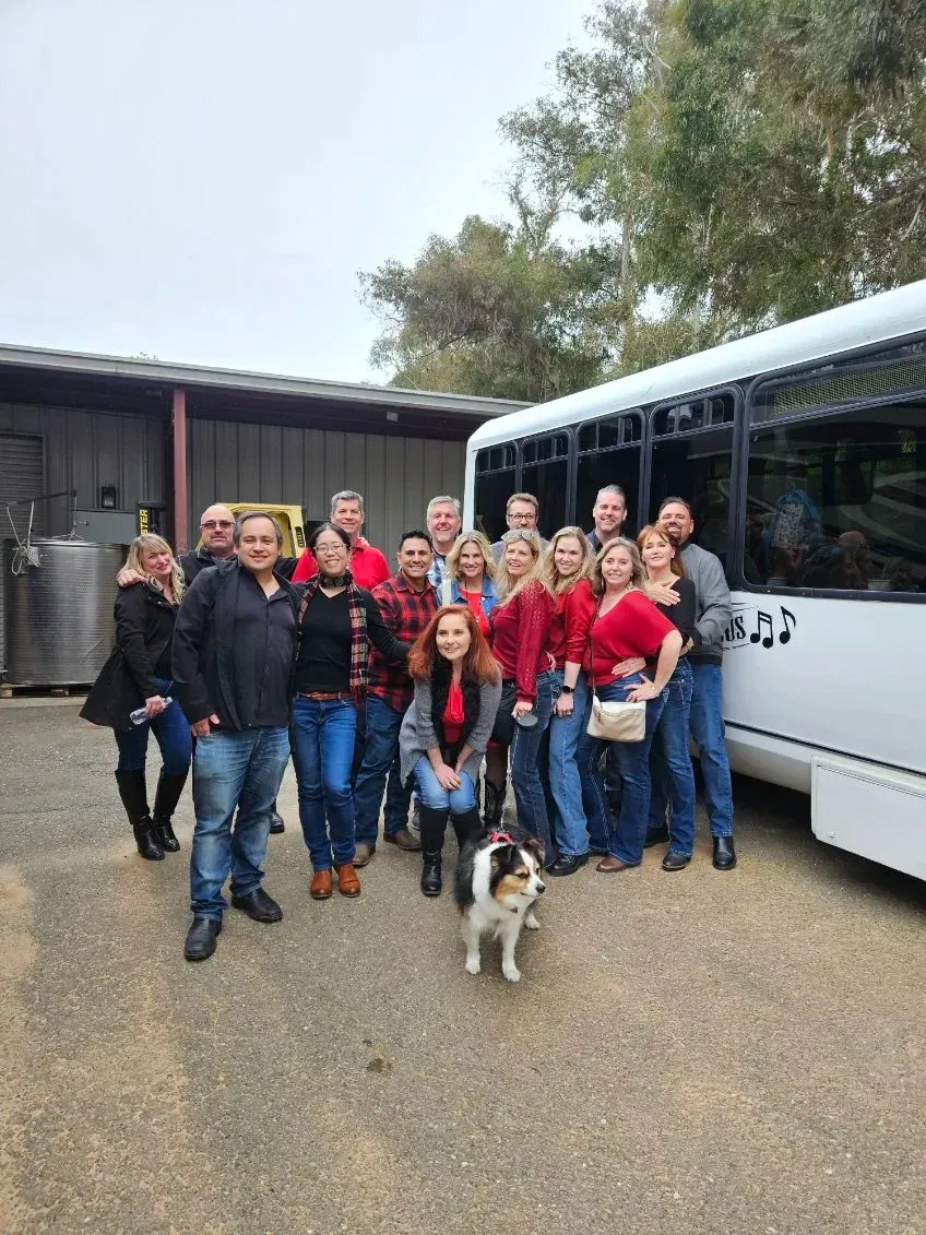 Group posing outdoors beside a white bus, with a small dog in front and buildings behind.