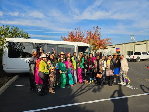 Group in colorful outfits posing beside a white bus in a parking lot on a sunny day