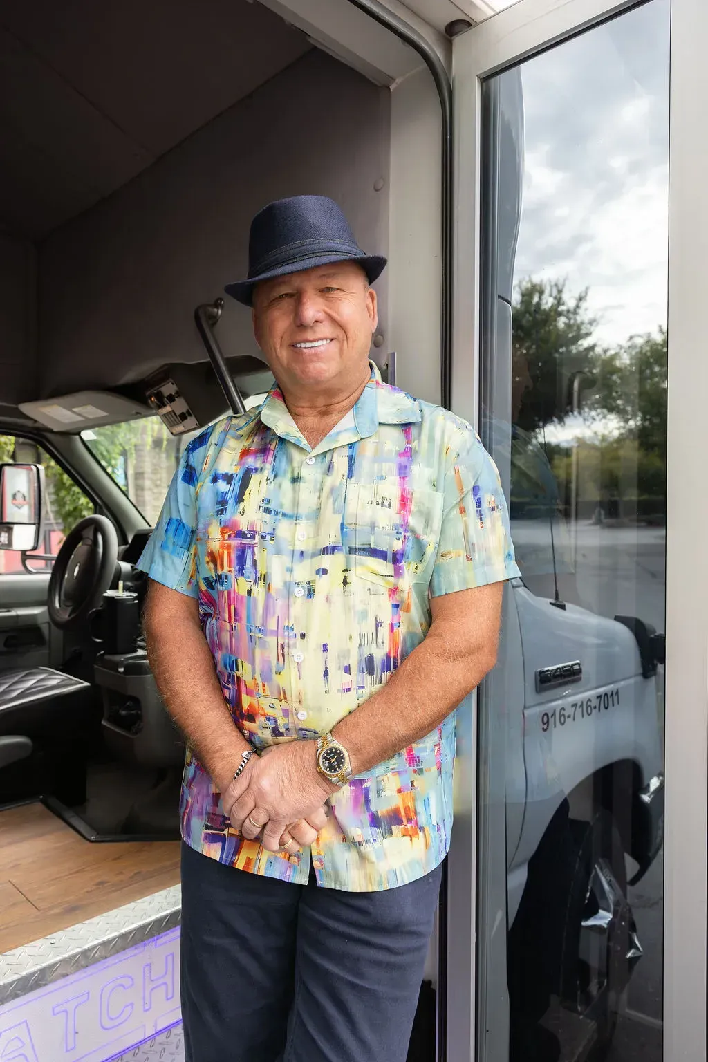 Man standing at a doorway, smiling, wearing a colorful shirt and hat beside a vehicle.