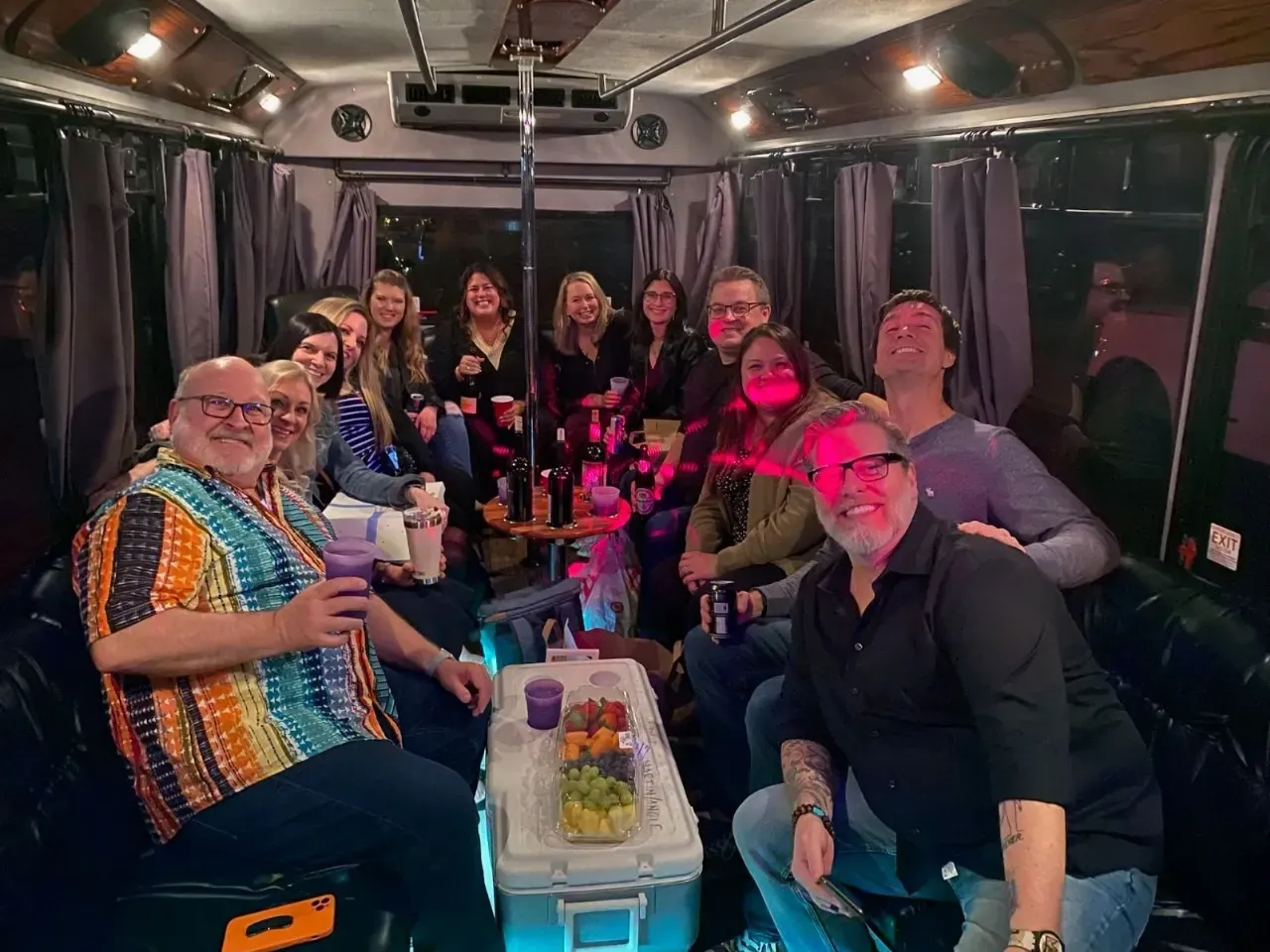 Group of people smiling in a party bus with pink lighting and a cake on the table.
