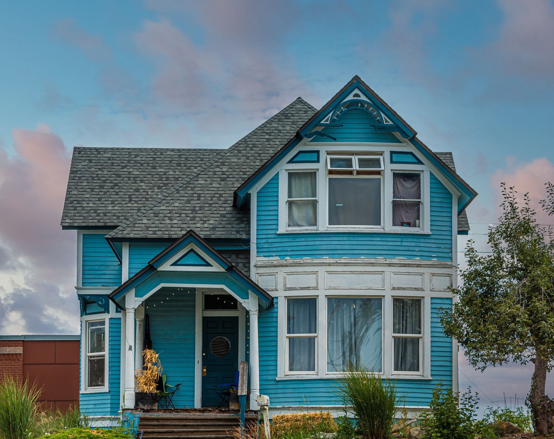 A weathered, two-story house with bright blue wooden siding and white trim under a soft, cloudy sky.