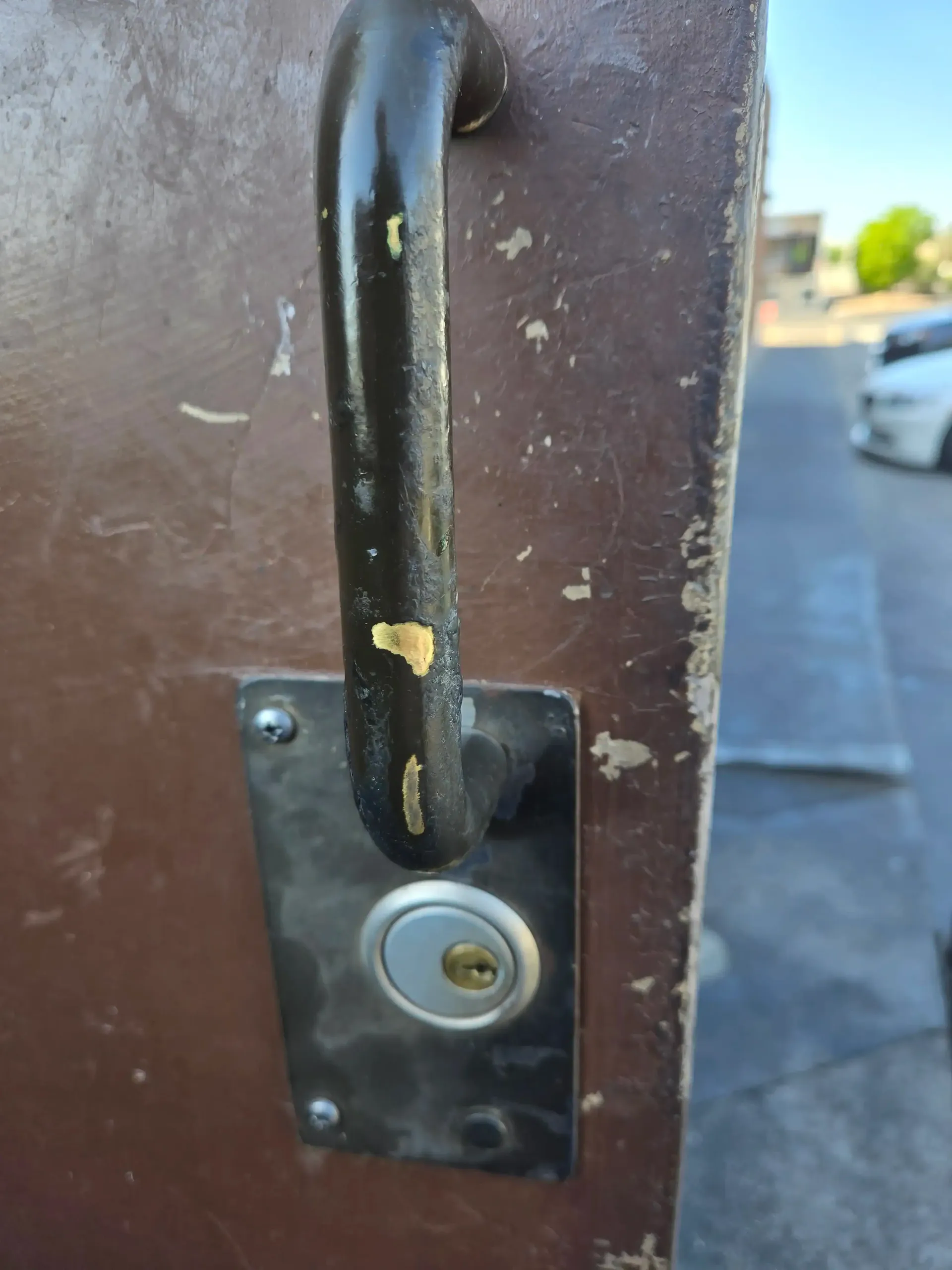 Close-up of a black metal handle and lock on a brown gate, with a street blurred in the background