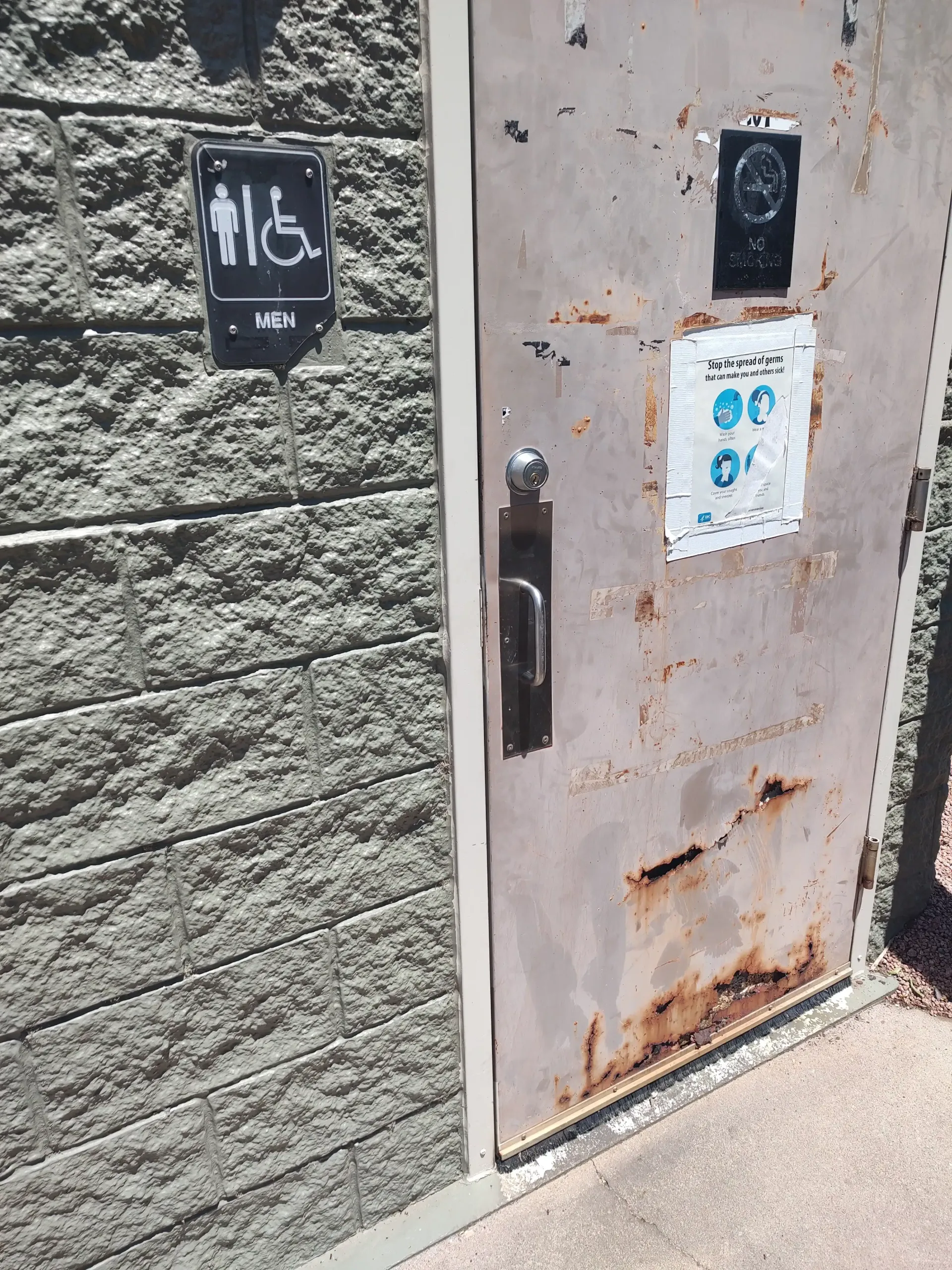 Rusty metal door with black handle and signs, set in a concrete wall beside a paved walkway.