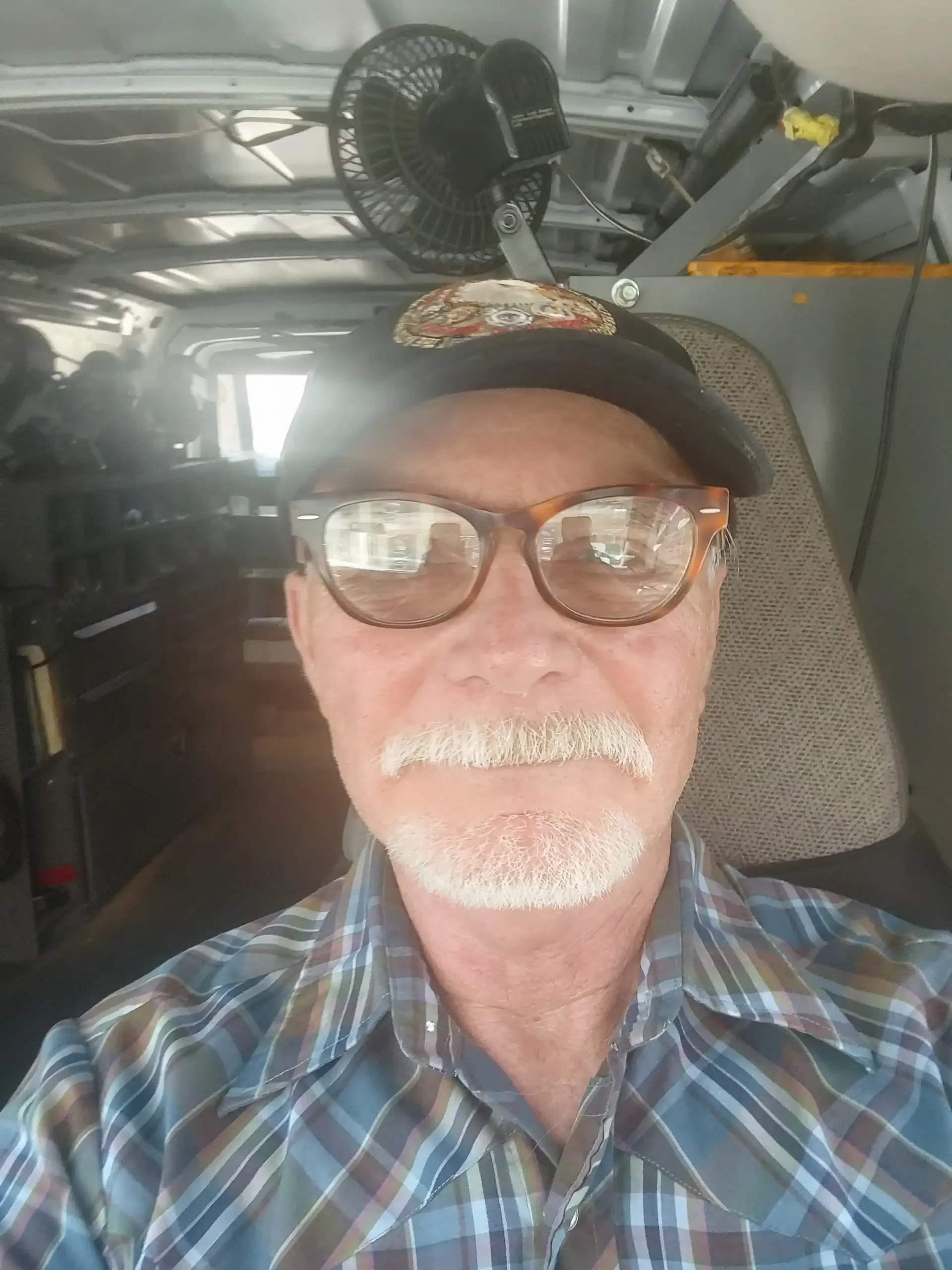 Man wearing glasses and a cap, seated inside a vehicle cab with a fan overhead.