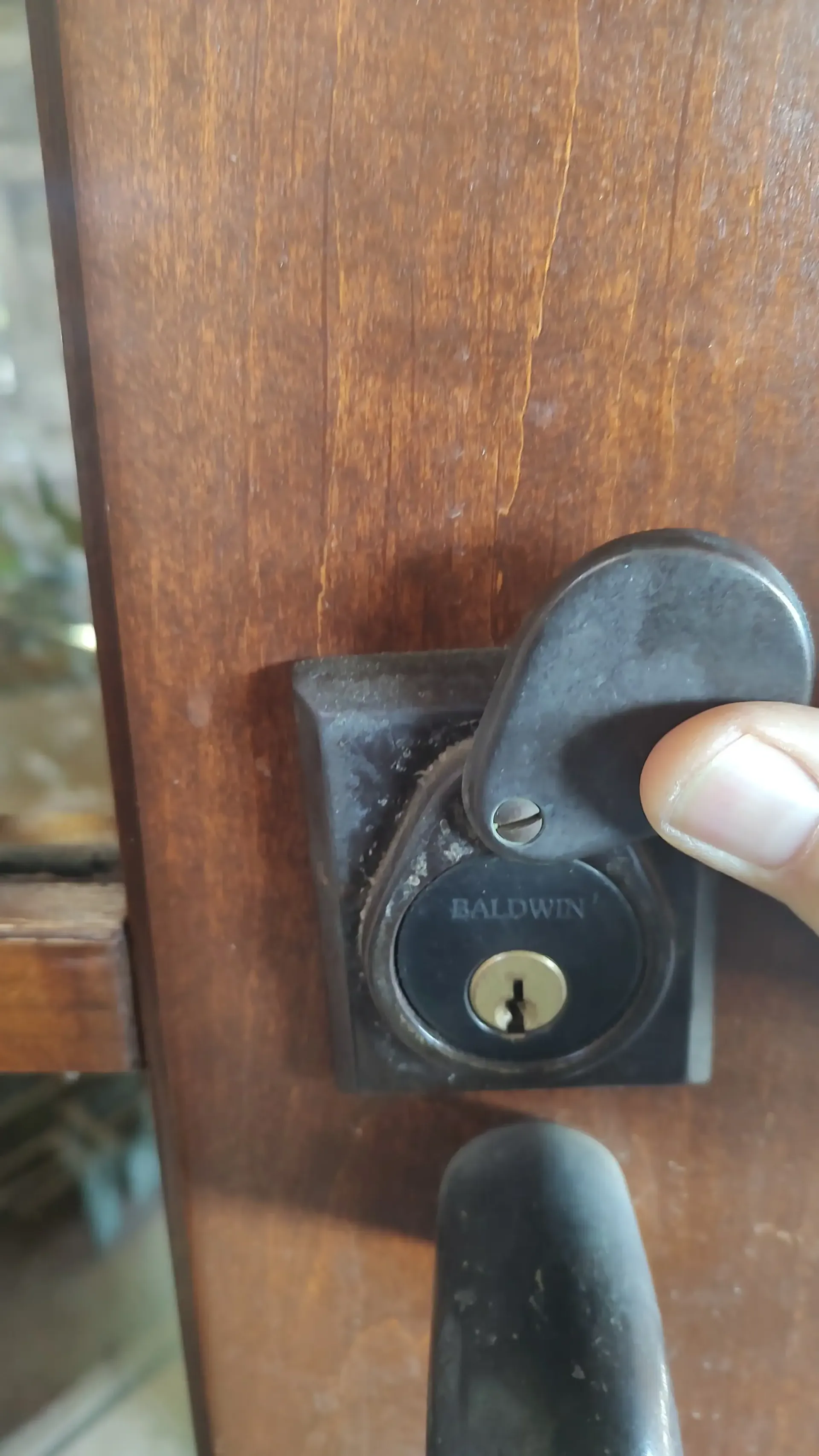 Finger unlocking a black deadbolt on a wooden door with a keyway and handle.