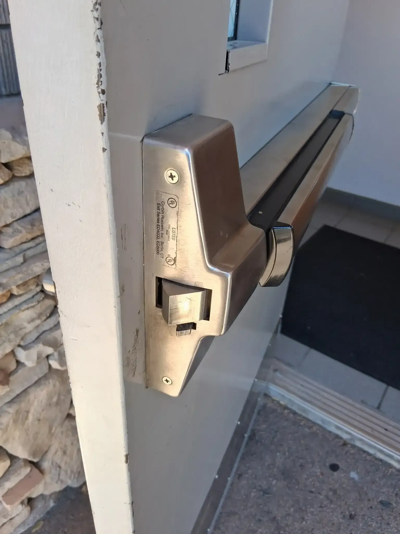 Close-up of a metal door exit bar and latch on a white wall by an outdoor step.