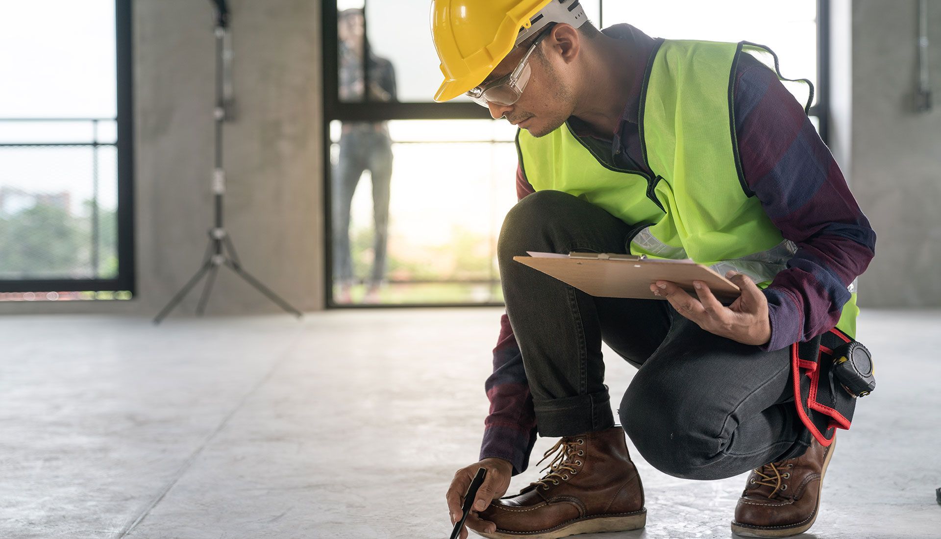 A construction worker is kneeling down and looking at a clipboard.