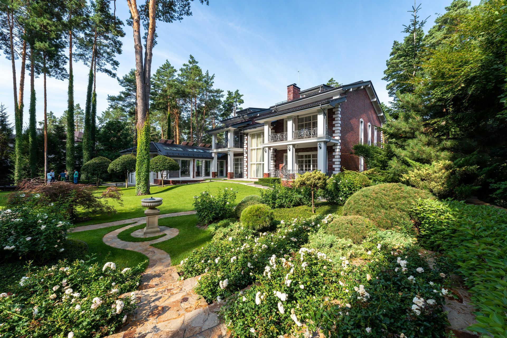 Manicured garden with a stately brick house, white columns, and a winding stone path under blue sky