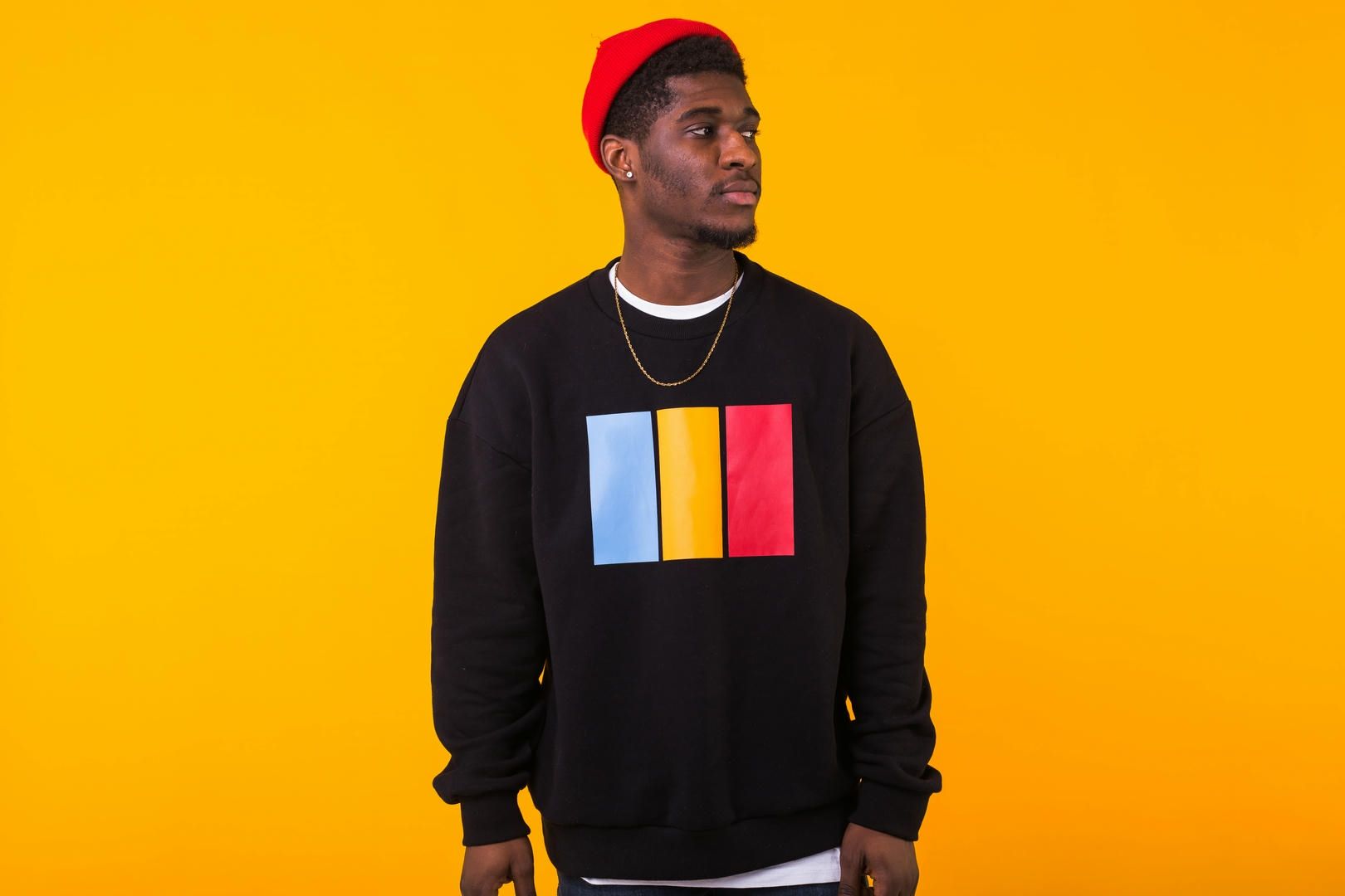 A man wearing a black sweatshirt and a red hat is standing in front of a yellow background.