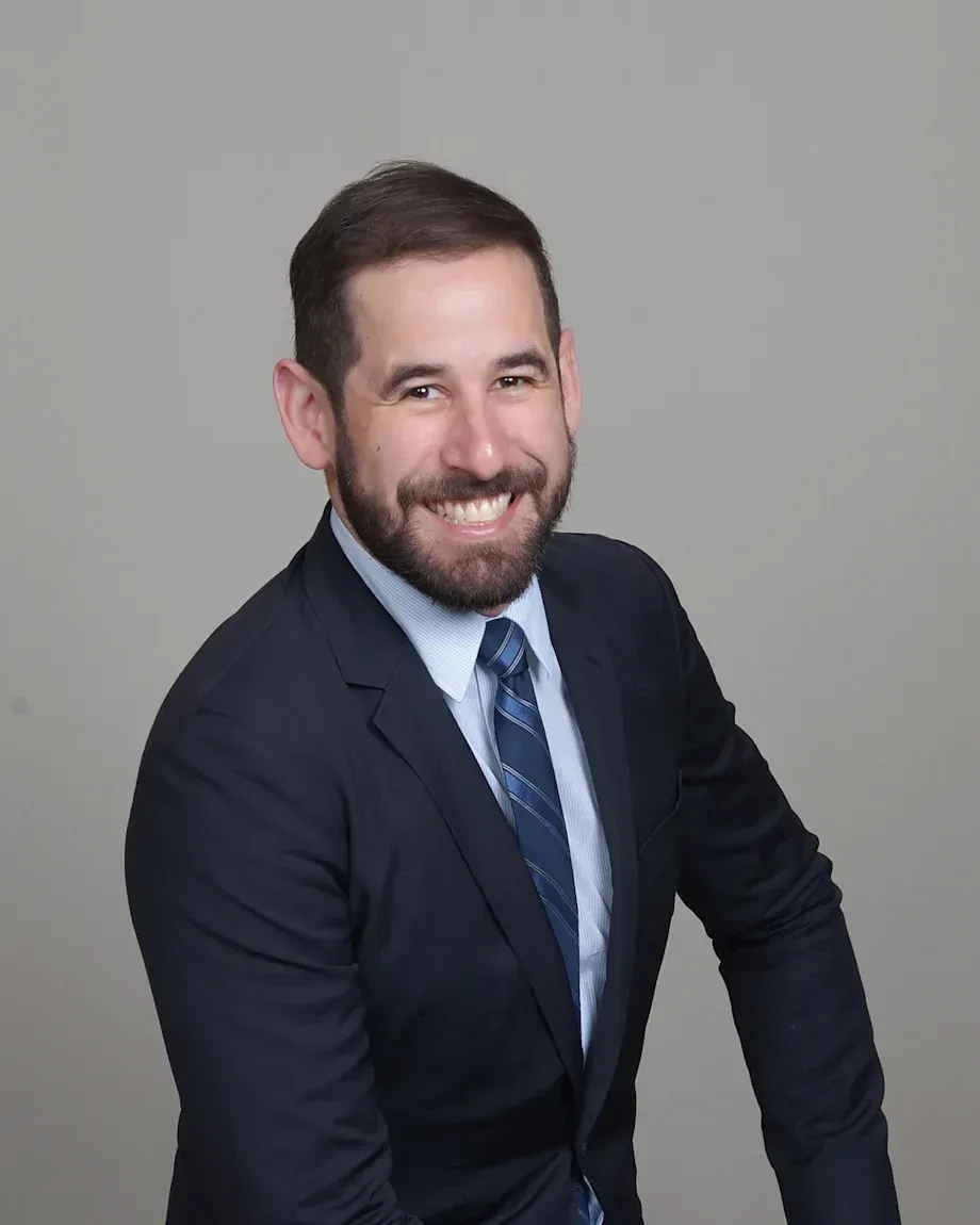 Man in a navy suit and tie, smiling, with a beard, against a gray background.