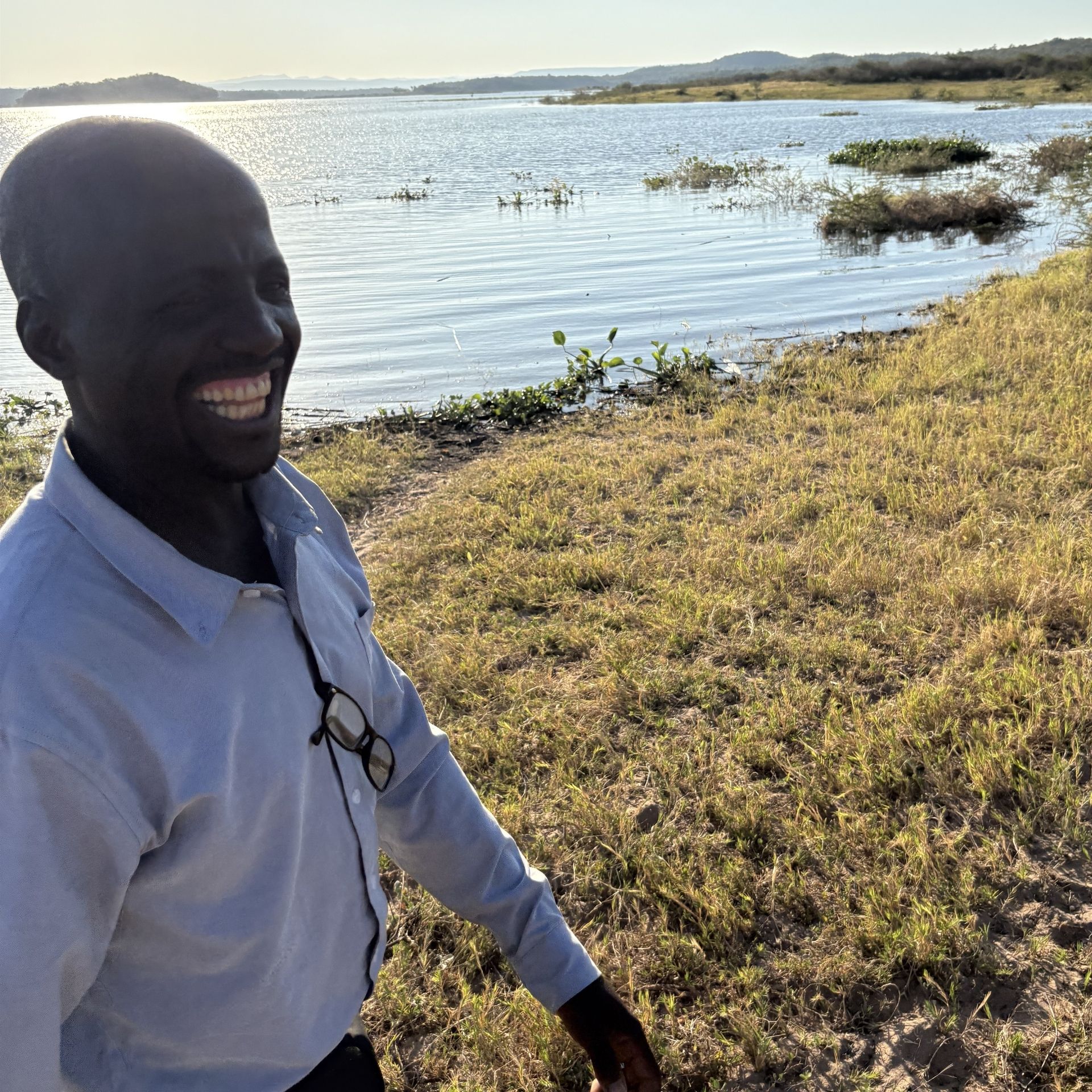 Smiling person in a light blue shirt by a lakeshore with birds on the water and grassy shoreline.