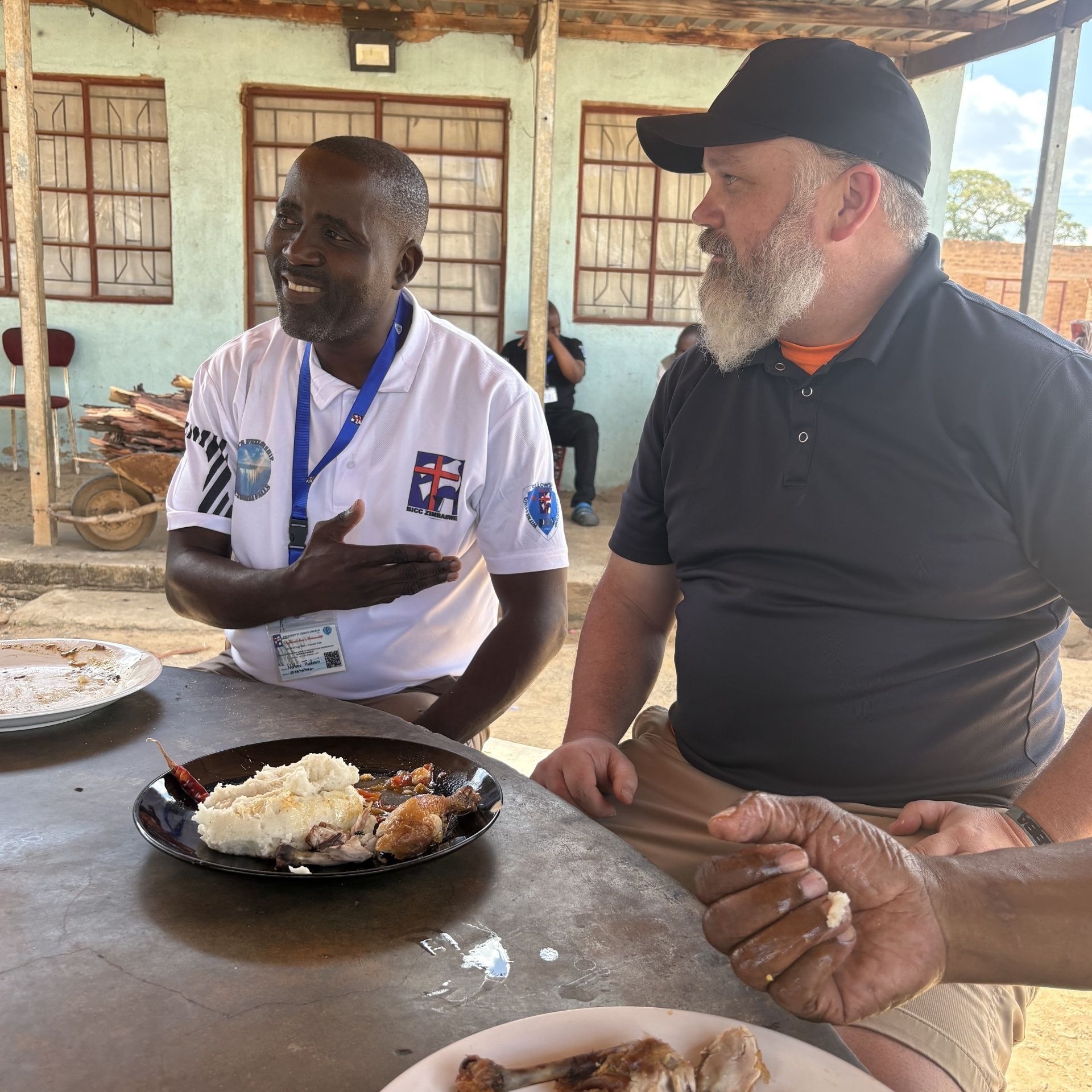 Two men sit at a table with plates of food in a casual outdoor dining area.