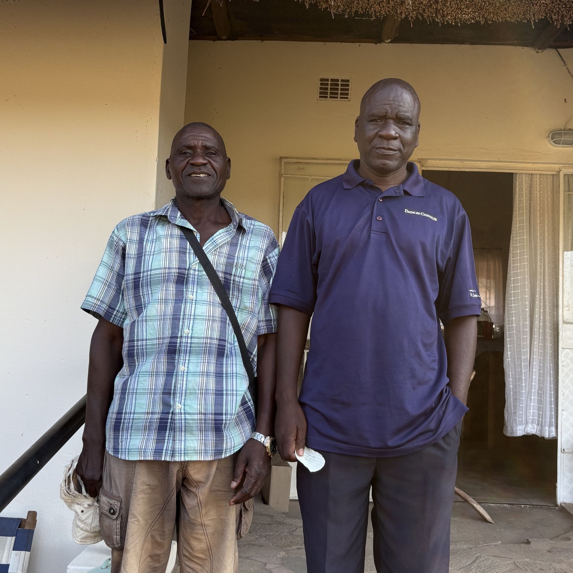 Two men standing outside a beige building, one in a plaid shirt and one in a navy polo shirt.