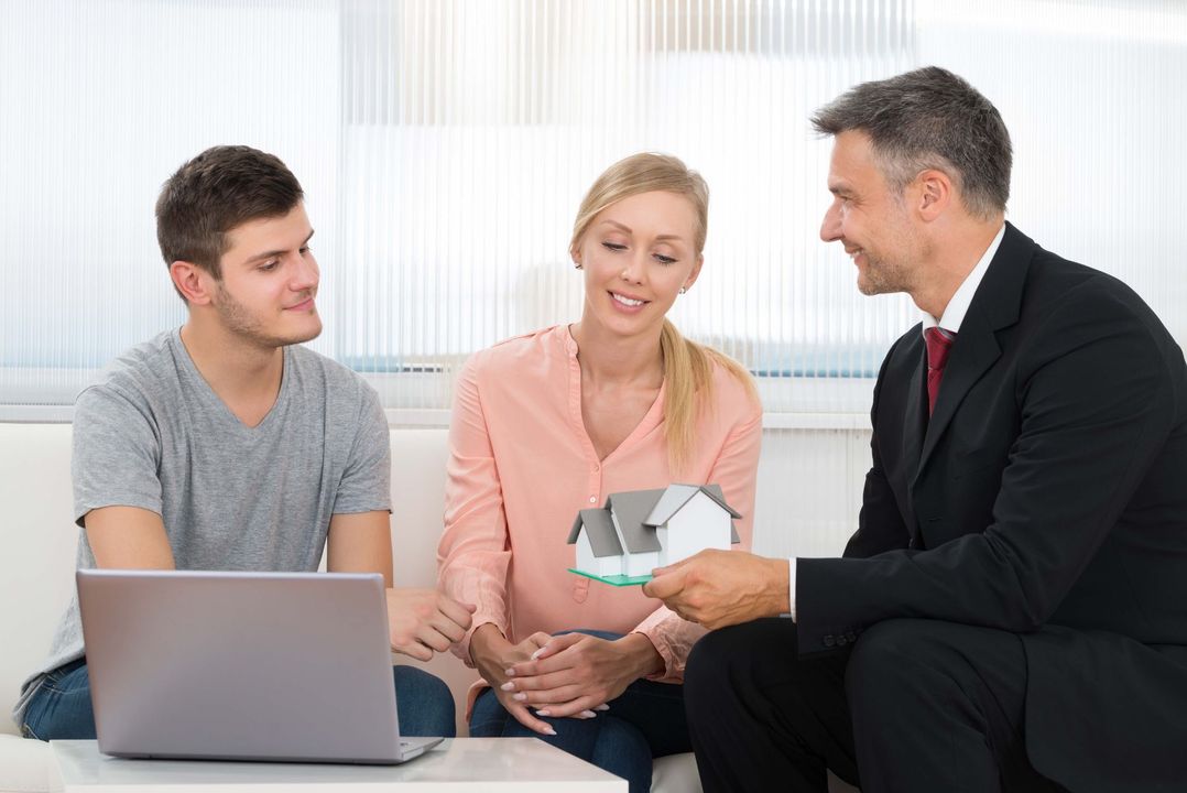 a man is sitting on a couch talking to a man and woman while holding a model house