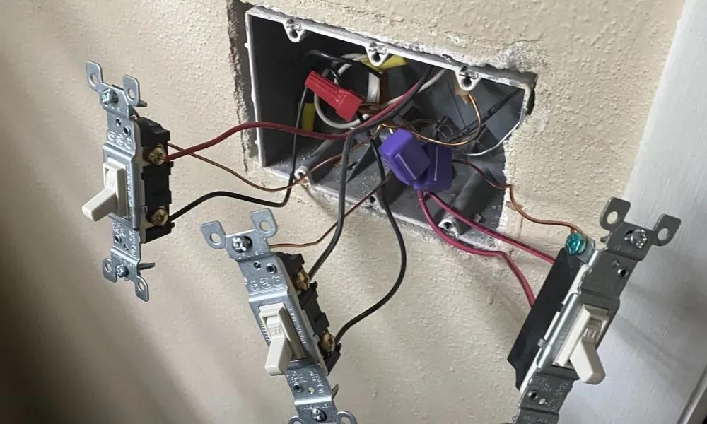 Three light switches, pulled from a wall-mounted electrical box, showing exposed wiring and wire connectors inside the wall.
