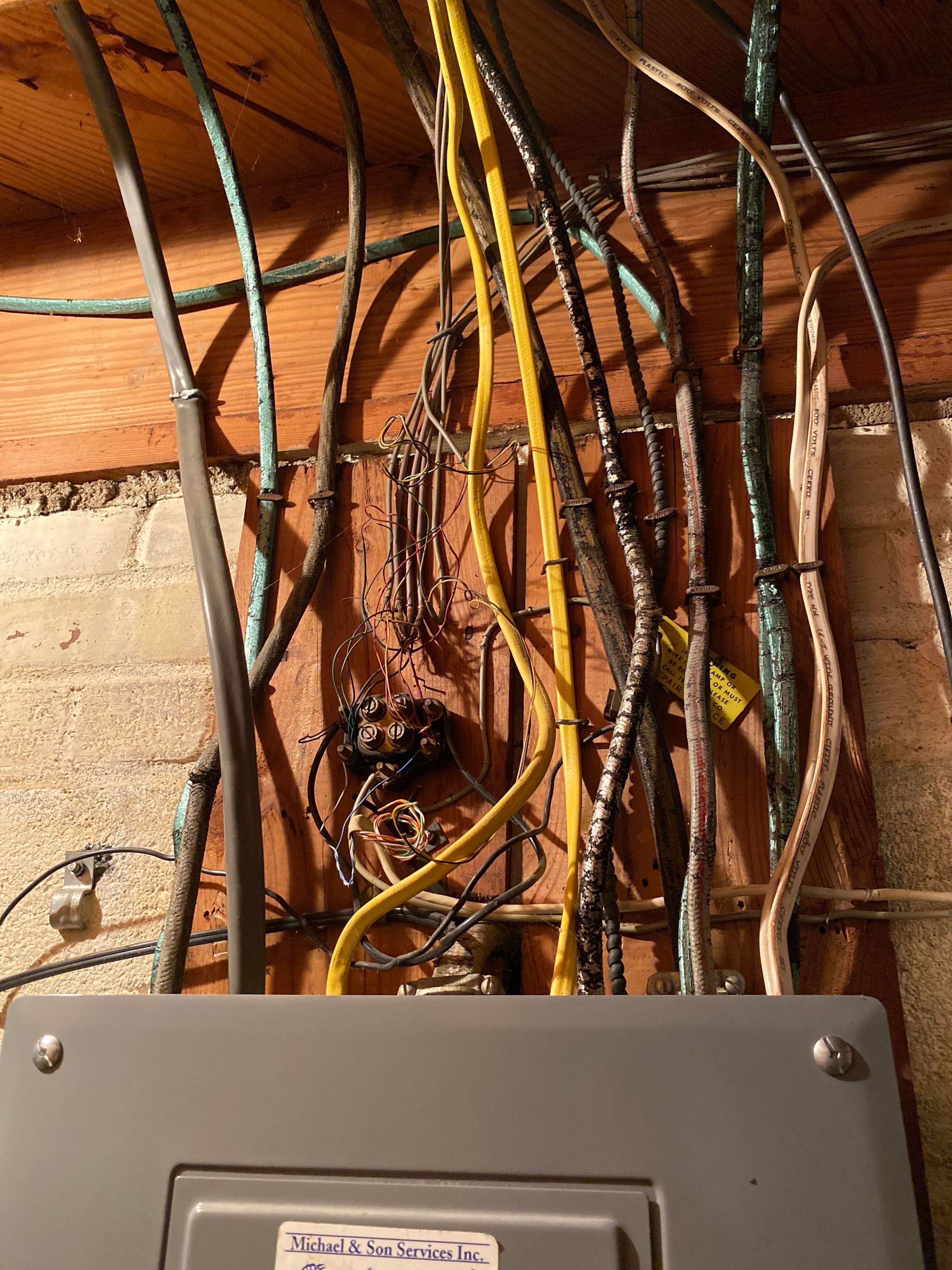 Electrical wires and cables running up a wooden wall and into the top of a metal circuit breaker box.