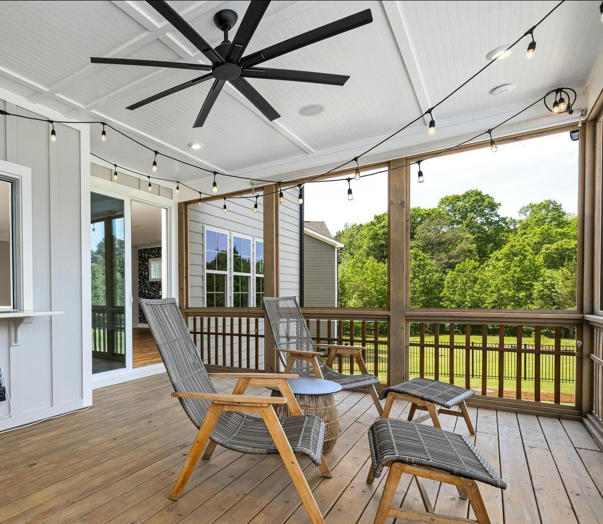Screened-in porch with seating, string lights, and a large ceiling fan; overlooking greenery.