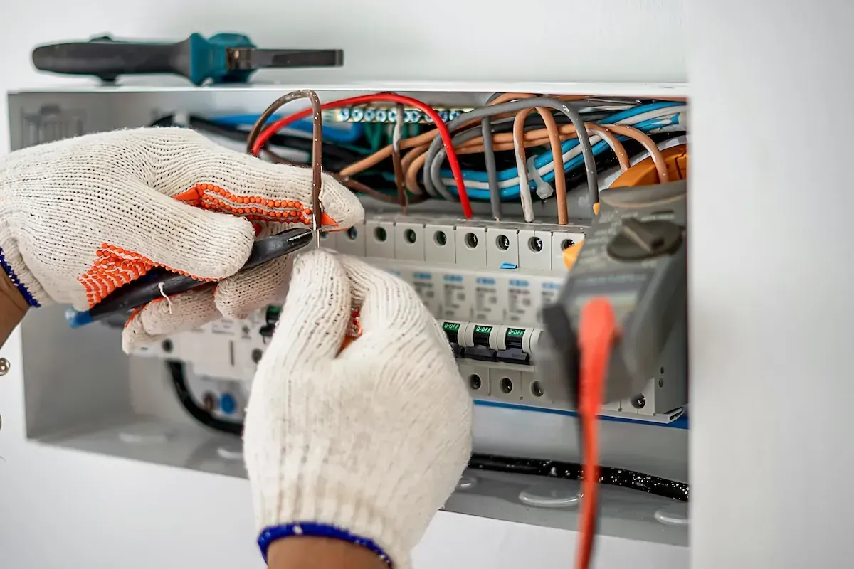 Hands in work gloves test electrical wires in a panel box with a multimeter.
