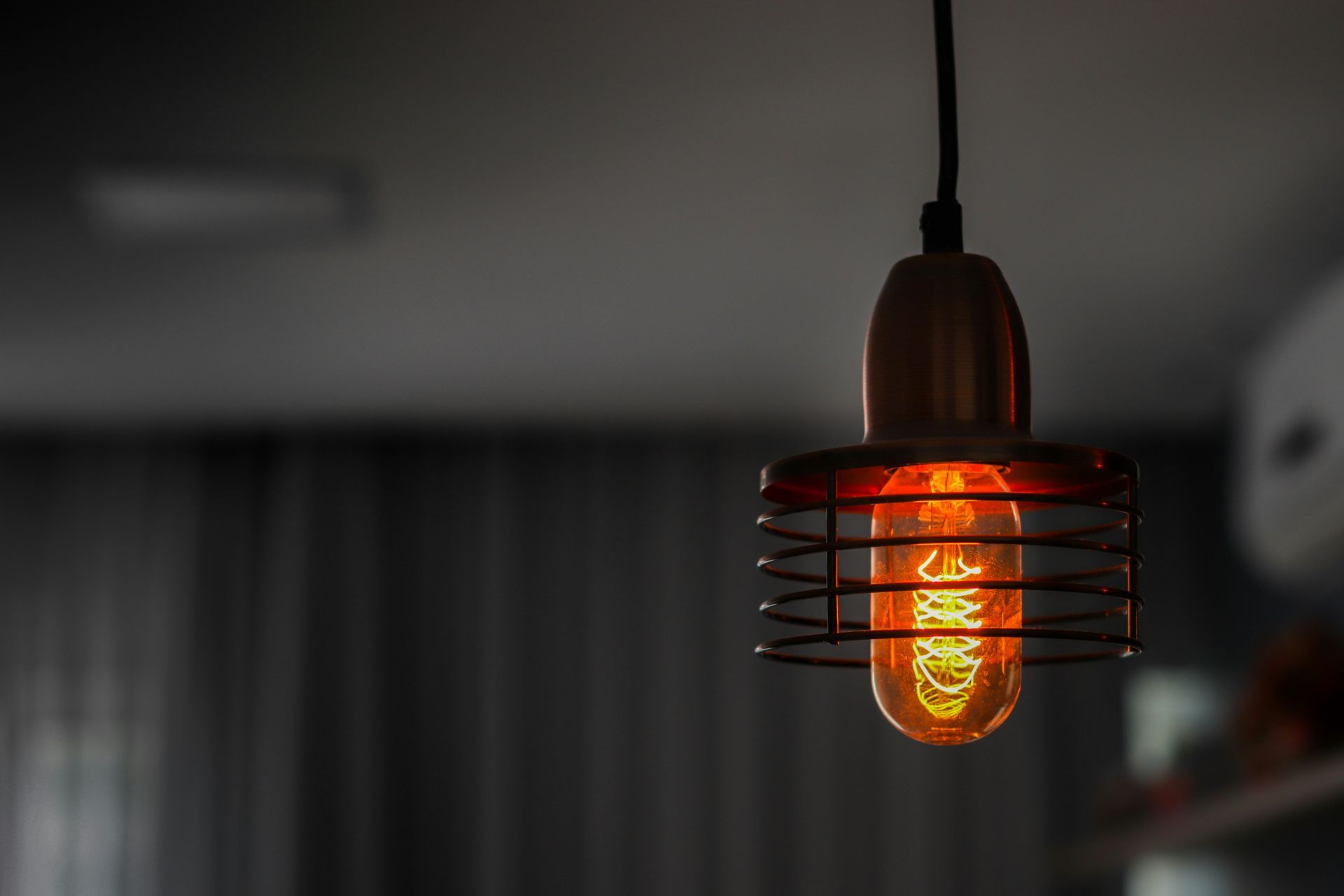 A glowing, amber-toned bulb hangs inside a decorative wire cage fixture against a blurred indoor background.