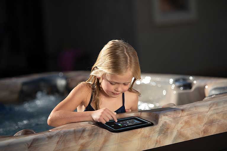 Girl in a hot tub, using a tablet. Brown tub surround with water and dark background.