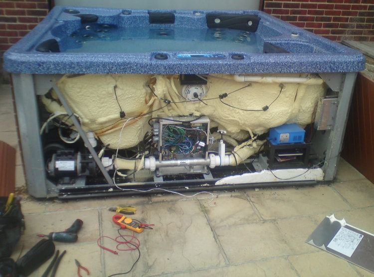 Hot tub, back panel removed, showing internal components. Electrical wires, pump, insulation, and repair tools visible.