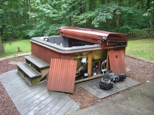 Hot tub with cover open, revealing the interior mechanics. Wooden steps and panels, outdoor setting.