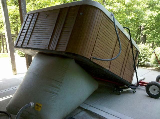 A hot tub tilted on a large, inflatable support, being moved on a dolly outdoors. Brown siding, green background.
