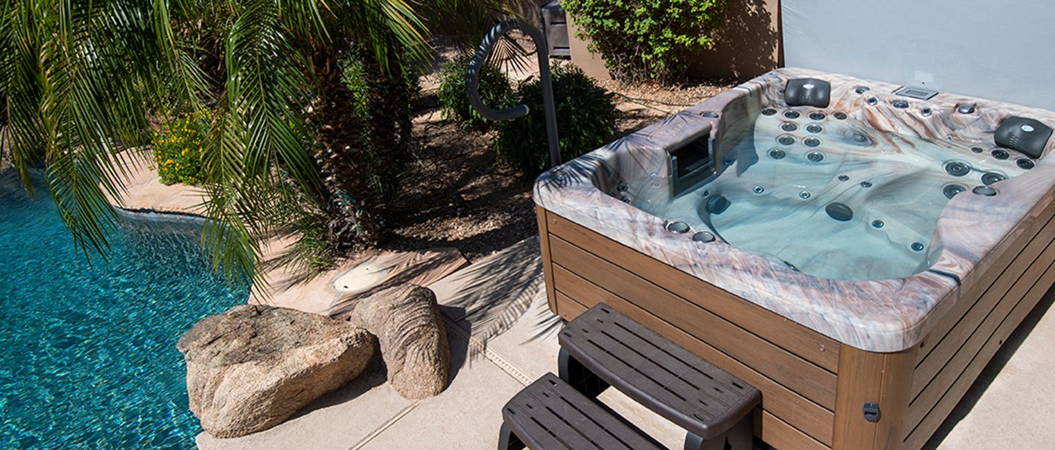 A hot tub next to a pool with steps, surrounded by rocks and foliage.