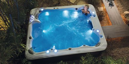 Person relaxing in a blue illuminated hot tub at night. Wooden walkway and greenery in the background.
