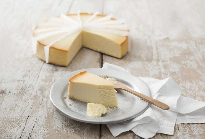 Una fetta di cheesecake su un piatto con una forchetta di legno e una cheesecake più grande sullo sfondo, su un tavolo di legno bianco.