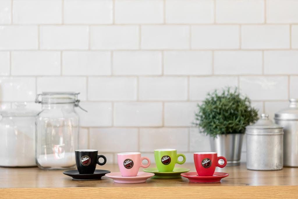 Quattro tazzine da caffè espresso colorate con relativi piattini su un bancone in legno, con uno sfondo di piastrelle bianche e una piccola pianta in vaso.