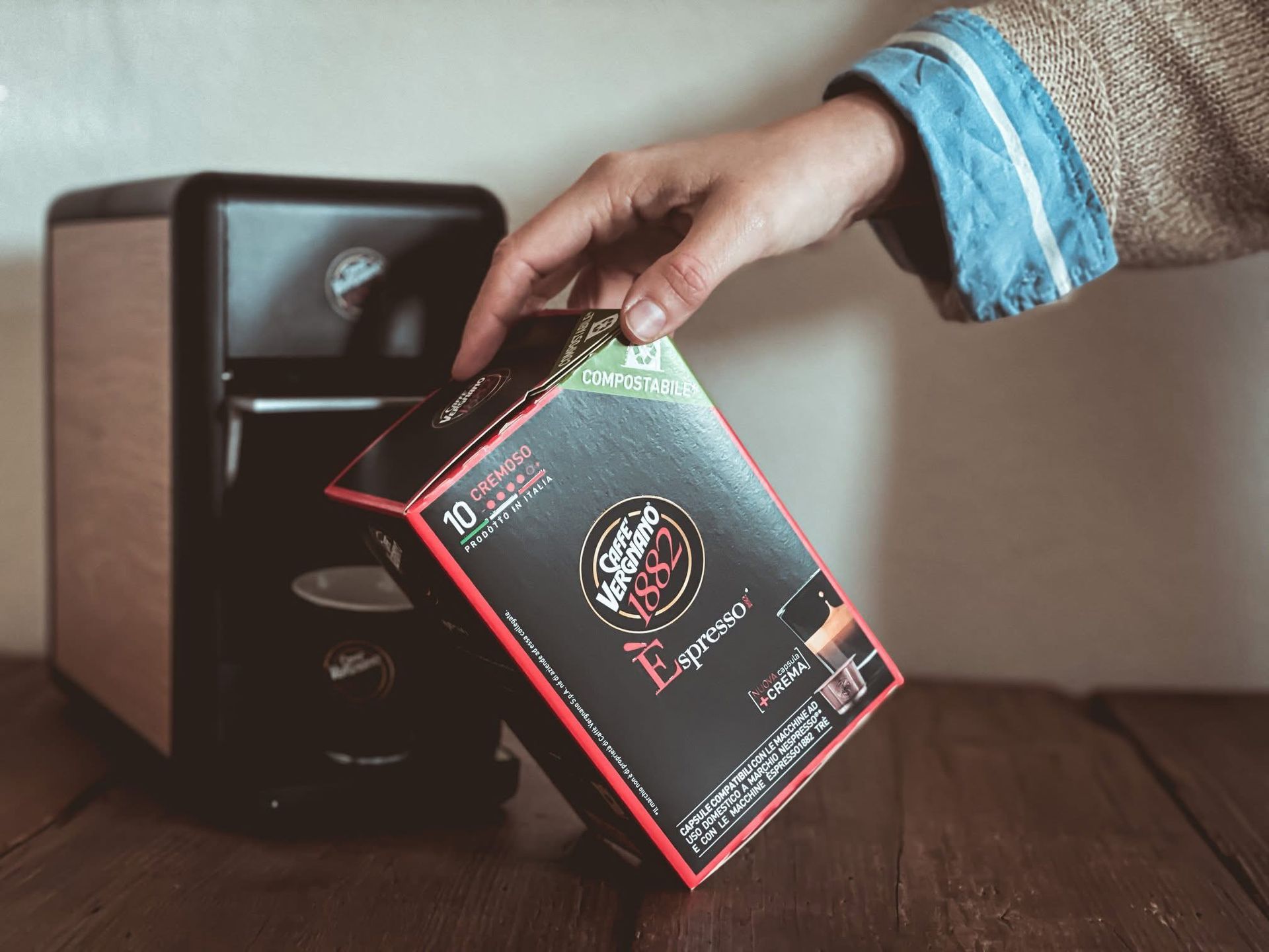Una mano che tiene una scatola di cialde di caffè nero vicino a una piccola macchina per caffè espresso nera e effetto legno, appoggiata su una superficie di legno.