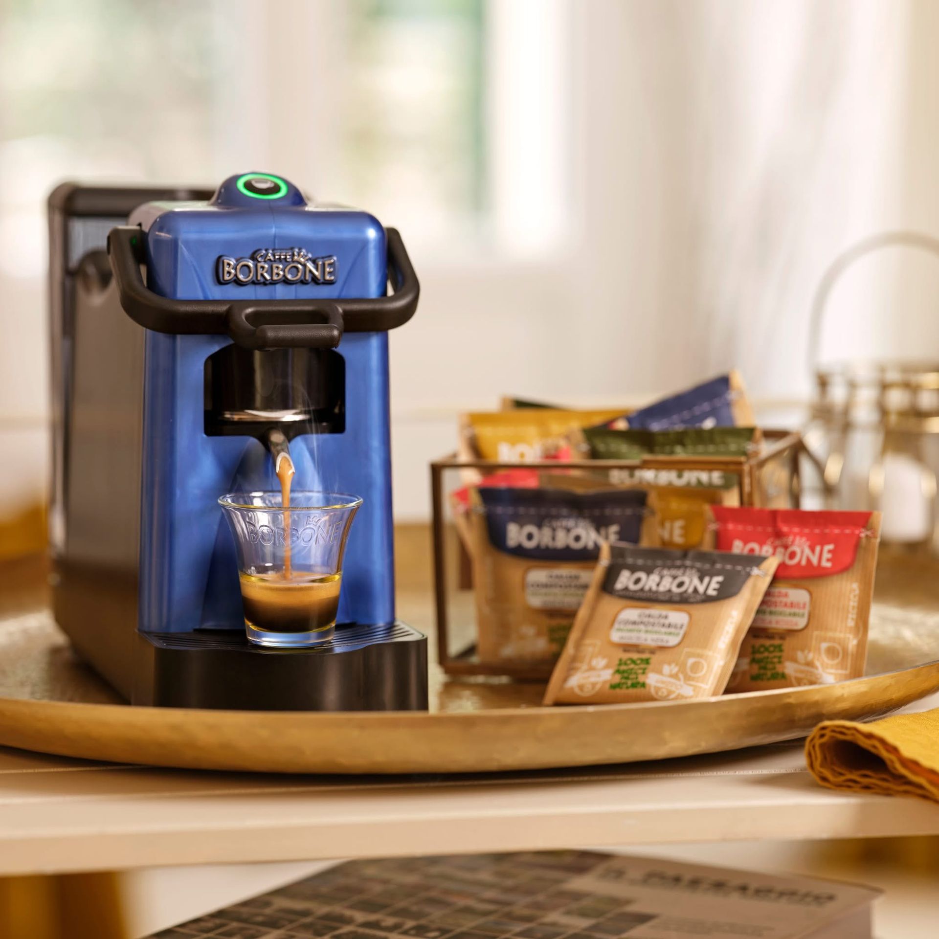 Macchina per caffè espresso blu che eroga il caffè in un bicchiere, con cialde e accessori su un vassoio.