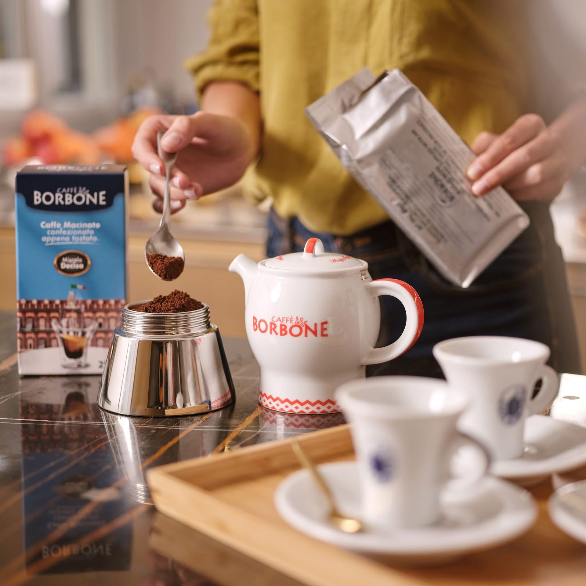 Una persona che prepara il caffè: versa il caffè macinato in una moka, vicino a una scatola di caffè Borbone, una teiera e delle tazze.