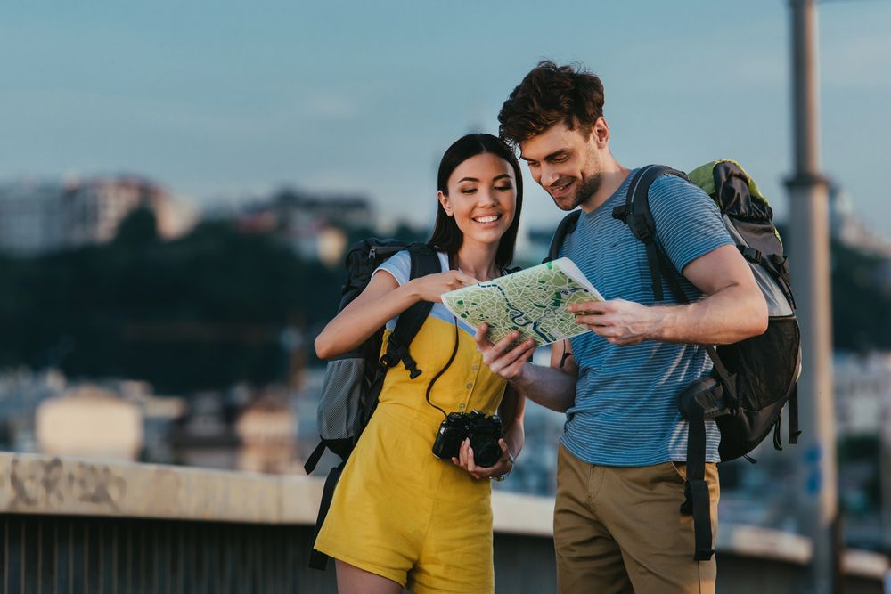 Pareja con mochilas mirando un mapa al aire libre, sonriendo. Ella lleva un mono amarillo y ella una camisa azul. La ciudad está de fondo.
