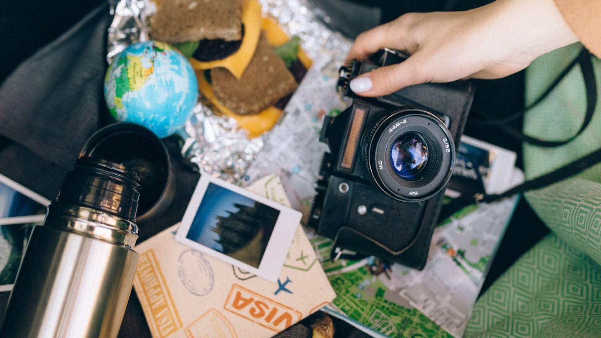 Mano sosteniendo una cámara negra, rodeada de artículos esenciales para el viaje: globo terráqueo, comida, termo, fotos, mapa.