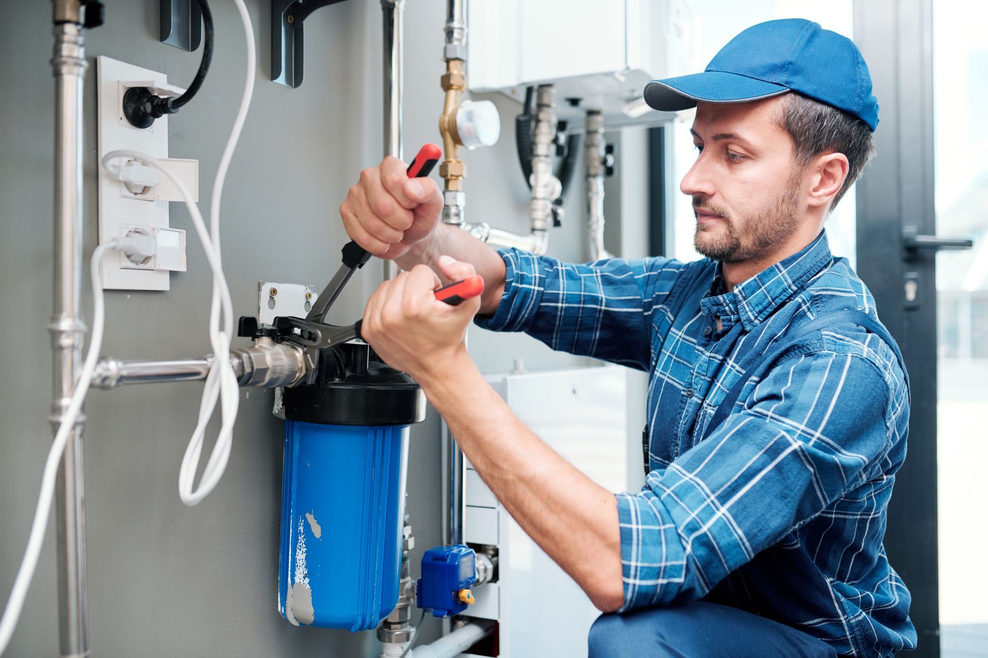 Plumber in a blue cap and plaid shirt working on water filtration system with tools. Plumber in a blue cap and plaid shirt working on water filtration system with tools.
