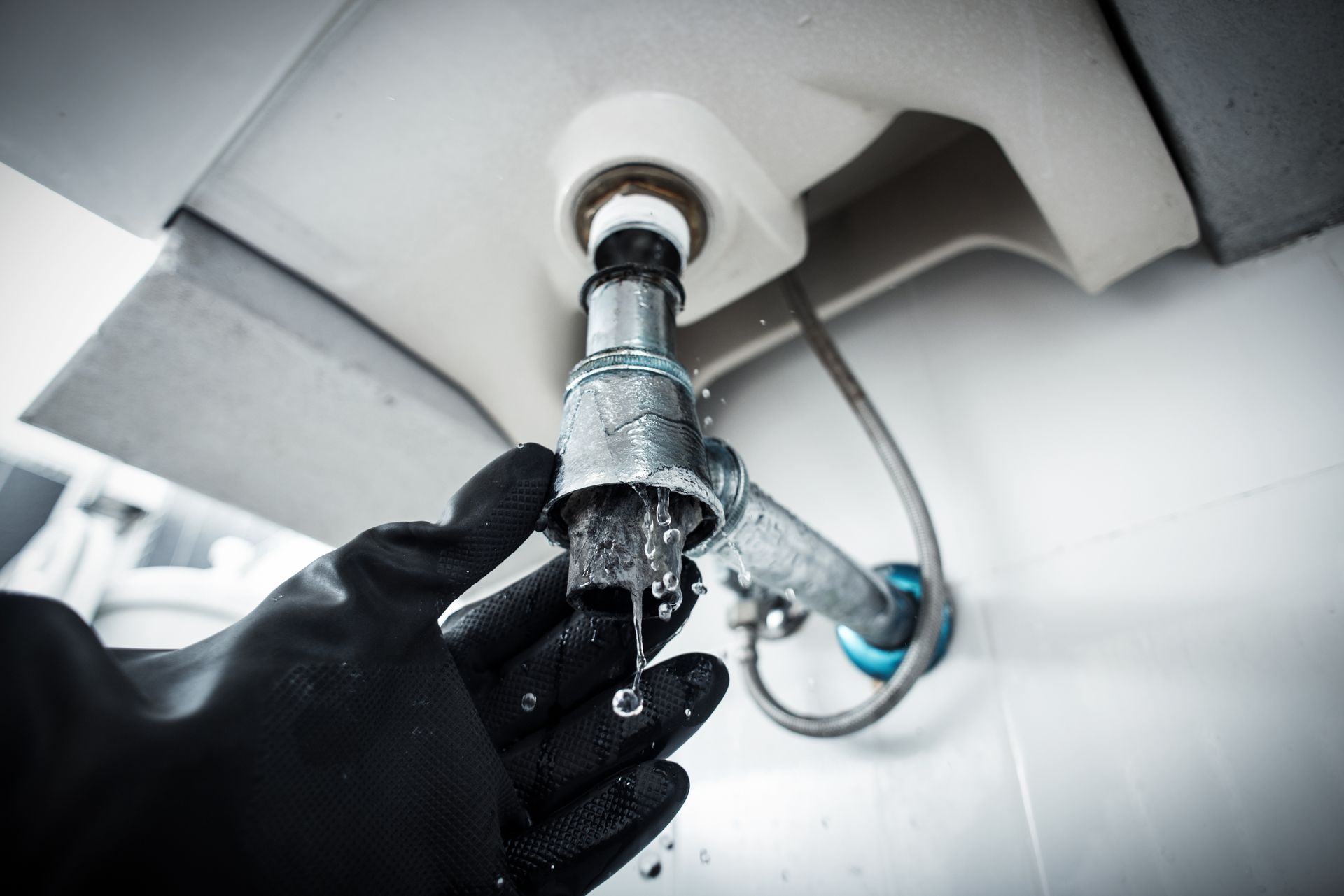 Hand in black glove tightening leaky sink pipe under a white sink.