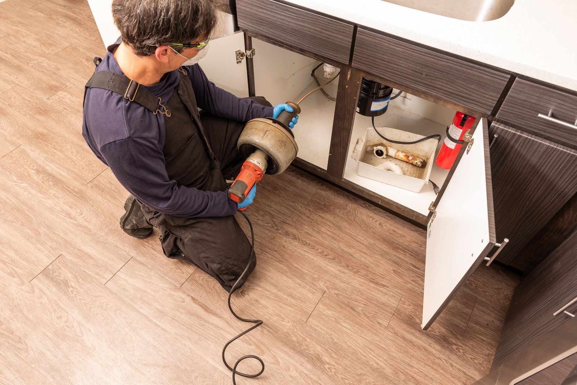 Plumber using a drain snake under a kitchen sink to unclog a drain.