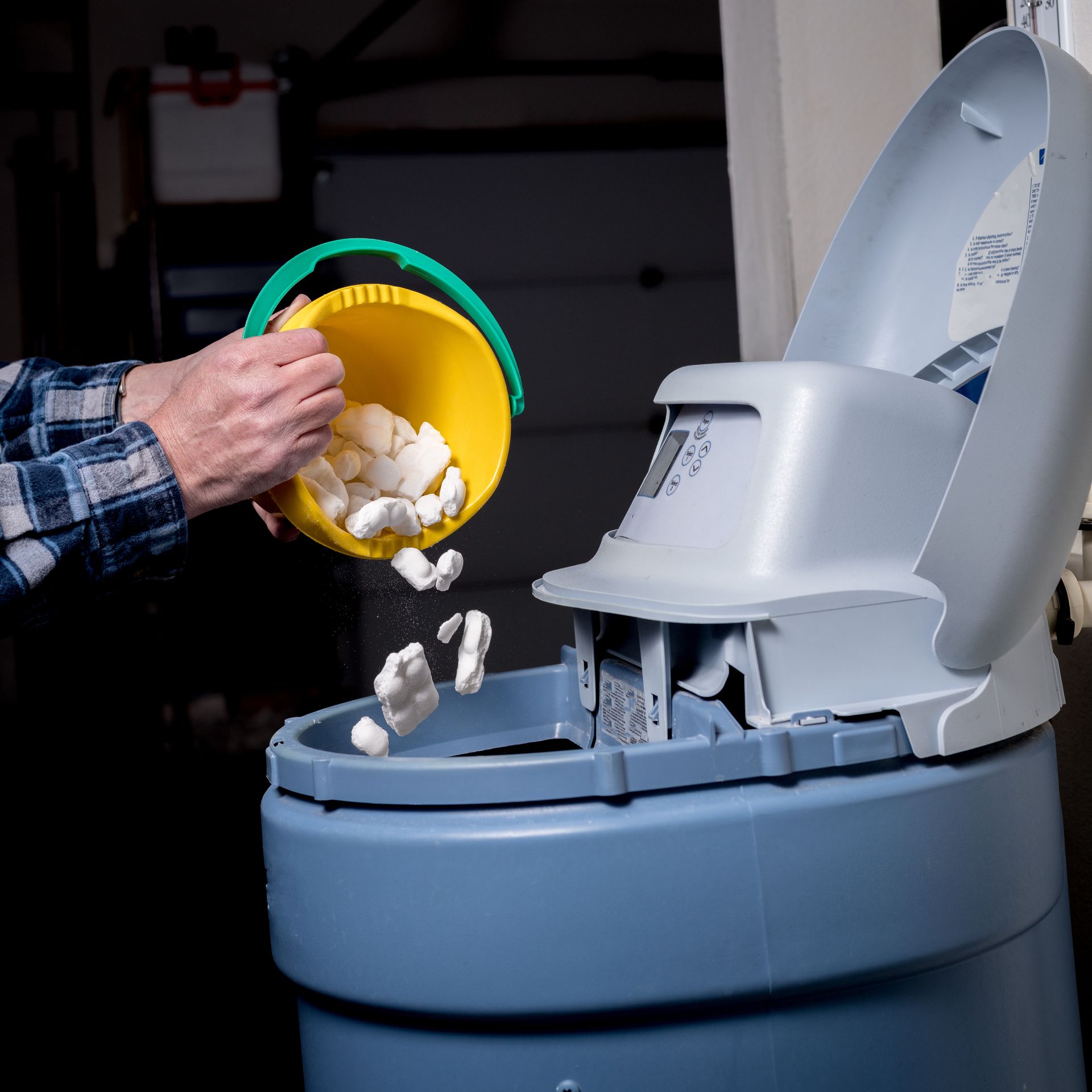 Person adding salt pellets to a blue water softener tank from a yellow bucket. Person adding salt pellets to a blue water softener tank from a yellow bucket.